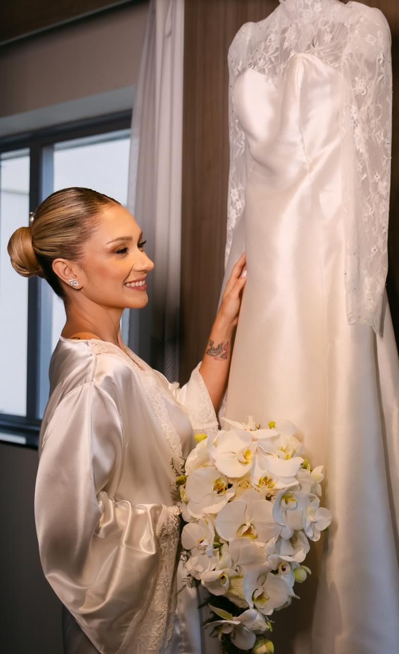 Noiva de robe de cetim sorrindo ao lado do vestido pendurado